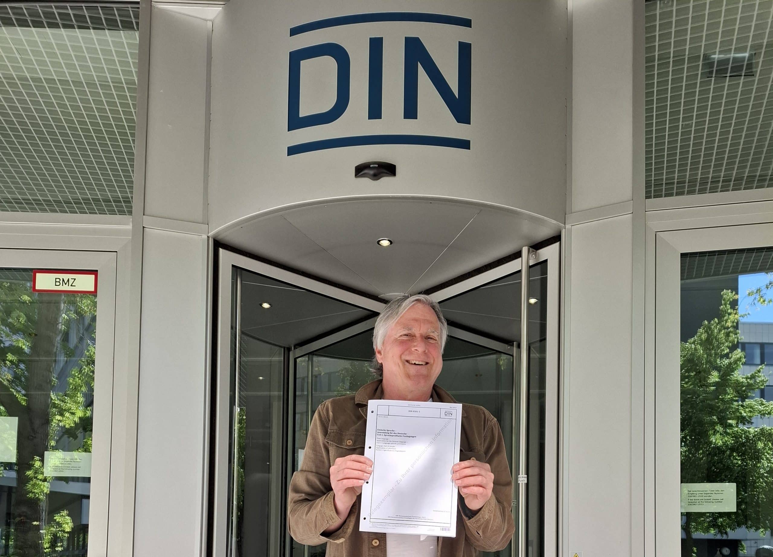 Uwe Roth steht vor dem Gebäude des Vereins Deutsches Institut für Normung. In der Hand hält er ein Exemplar der DIN 8581-1 Einfache Sprache. Foto: privat